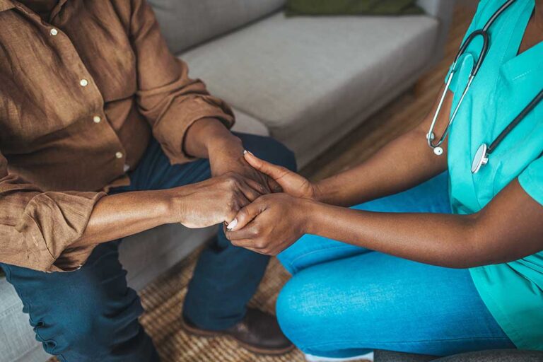 A middle-aged caregiver sits quietly beside an elderly parent, holding hands in a peaceful home setting.