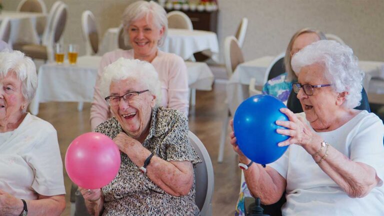 Older adults and children sit together at a table making colorful crafts in a bright activity room.